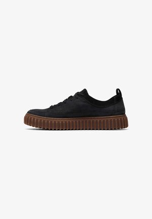 Zapatilla de ante negro con una suela de goma marrón texturizada, que presenta un perfil bajo, cordones redondos y una pequeña pestaña en el talón en la parte posterior.