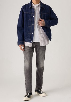 Mann trägt eine dunkelblaue Jeansjacke über einem weiß gestreiften Hemd, graue Jeans und schwarze Sneakers, steht vor einem schlichten Hintergrund.