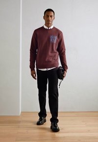 Felpa bordeaux con grafica stampata, camicia bianca a bottoni sotto, pantaloni neri e scarpe nere, mentre tiene una borsa piccola.