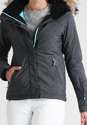 Kobieta ubrana w ciemnoszarą bluzę na suwak z jasnoniebieskim zamkiem i białe spodnie, pozująca z jedną ręką w kieszeni.