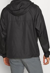 Man in een zwarte hoodie en grijze broek, van achteren te zien tegen een witte achtergrond.