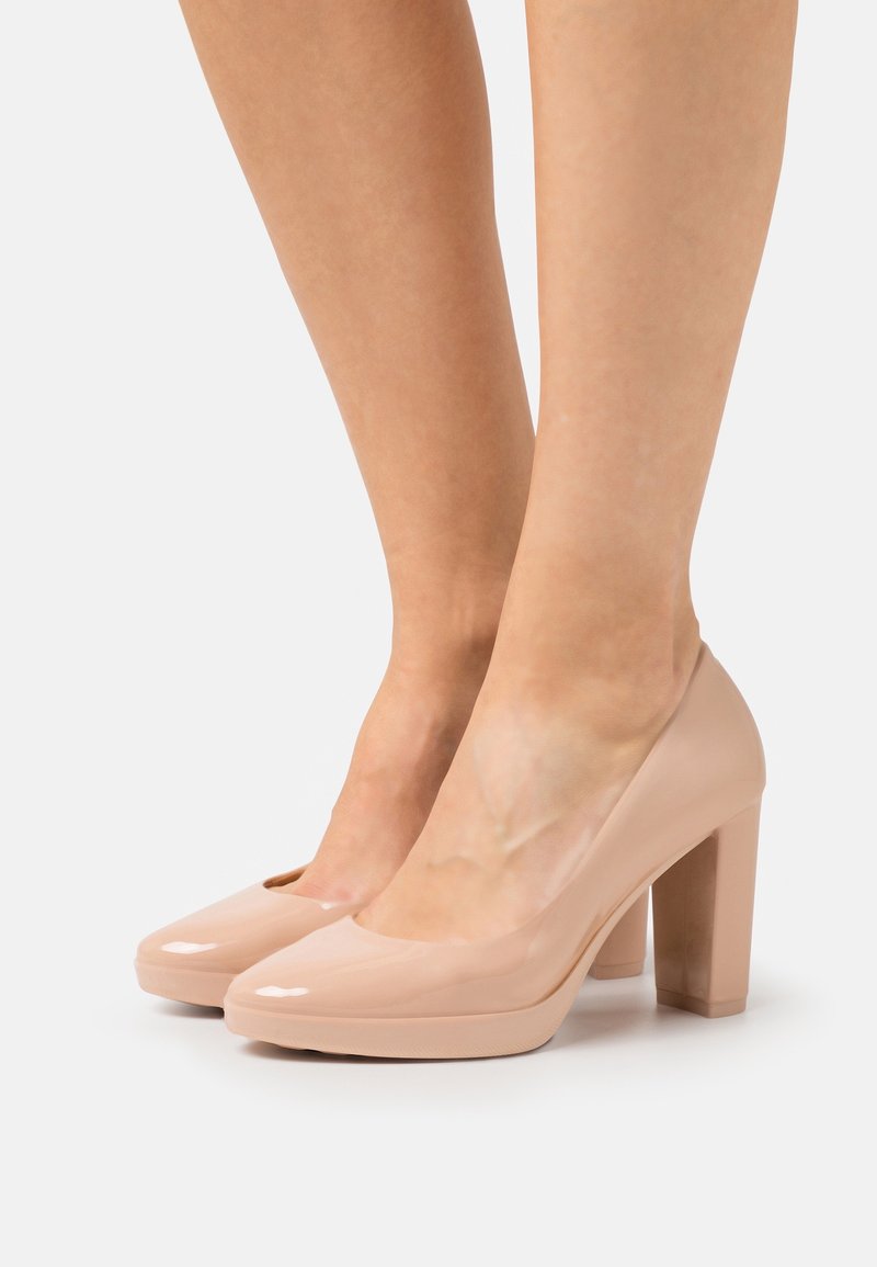 Jambes portant des escarpins en cuir verni nude brillants à talons blocs avec bouts arrondis sur fond blanc.