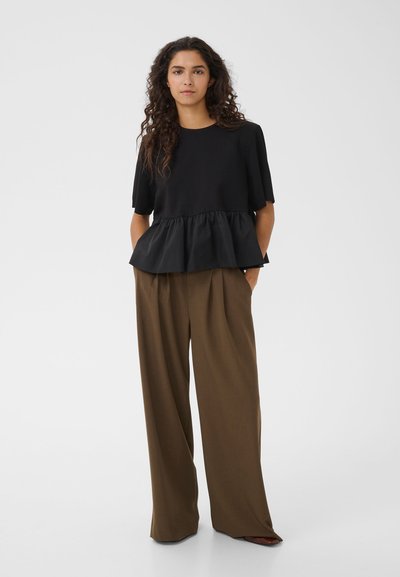 Femme aux longs cheveux bouclés portant un haut noir à manches courtes avec basque et un pantalon marron à jambes larges, debout les mains dans les poches.