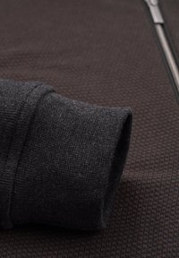 Schwarze Zip-Jacke mit strukturiertem Muster. Verfügt über eine dicke, gerollte Manschette aus einem einfarbigen schwarzen Material auf einem braunen Hintergrund.