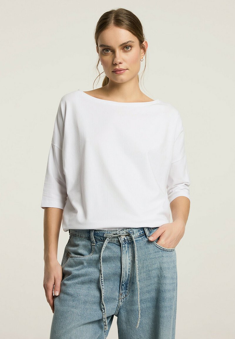 Biała, luźna bluzka z trzema ćwiartkami rękawów, wykonana z miękkiej bawełny; w zestawie z jasnodemnowymi dżinsami z wysokim stanem.