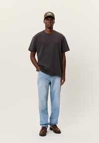 Σκούρο γκρι κοντομάνικο μπλουζάκι με μικρό λογότυπο, συνδυασμένο με ανοιχτό μπλε τζιν παντελόνι ίσιας γραμμής και καφέ παπούτσια με κορδόνια, ολοκληρωμένο με πράσινο καπέλο.