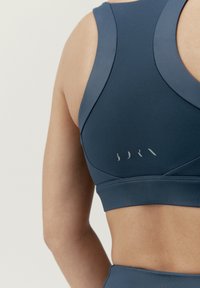 Tmavě modrá sportovní podprsenka s hladkým povrchem, širokými ramínky a minimalistickým logem na zadní straně. Tvarově přizpůsobený design.