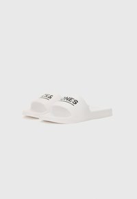 Sandales blanches à semelle plate avec une finition lisse et mate. Texte noir indiquant "JACK & JONES" sur la sangle supérieure. Design ouvert au niveau des orteils.