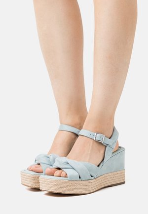 Pieds portant des sandales compensées en daim bleu clair avec des brides à la cheville et des semelles en corde tressée sur un fond uni.