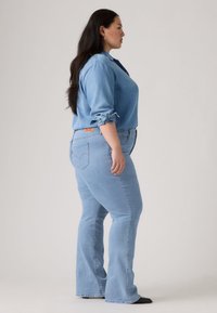 Svijetloplave traperice s uskim strukom i širokim nogavicama, uparene s odgovarajućom svijetloplavom traper košuljom, s uvrnutim rukavima i detaljima na gumbima.