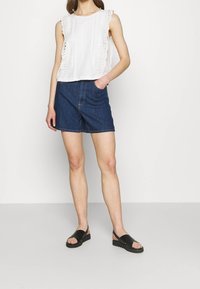 Vit ärmlös topp med texturerad spetsdetalj, i kombination med mörka denimshorts och svarta platta sandaler. En enkel och avslappnad outfitdesign.