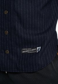 Marineblå skjorte med nålestriber og guldtonede knapper. Har en mærkat med teksten "Mitchell & Ness" samt "Sporting Goods" og stednavn.