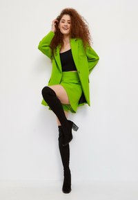 Ensemble blazer et jupe verts avec un crop top noir ; bottes hautes en suède noir avec un talon carré ; boucles texturées ; fond blanc simple.