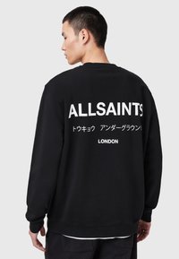 Man facing sideways wearing a black sweatshirt with "ALLSAINTS," Japanese text, and "LONDON" printed in white on the back.