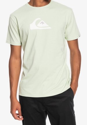 Man wearing light green short-sleeve t-shirt with white mountain wave logo on chest and black pants, standing with one hand in pocket.