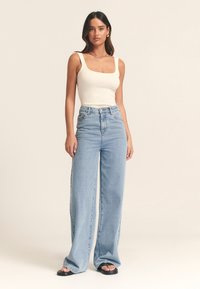 Gai bēšs ribots tanks saskaņots ar gaiši ziliem augsta vidukļa, platajiem džinsa džinsiem. Modelis nēsā melnas plakanas sandales.