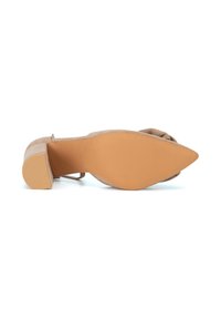 Zapato de tacón alto de ante beige con punta puntiaguda, textura lisa y suela de goma marrón claro. Presenta un detalle frontal anudado.