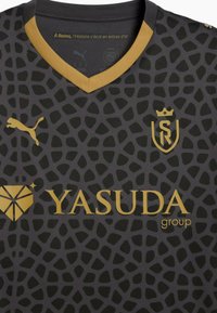 Sort fodboldtrøje med et struktureret mønster i mørkegrå, udstyret med en guld V-udskæring og guld branding: Puma-logo og Yasuda-gruppe tekst.