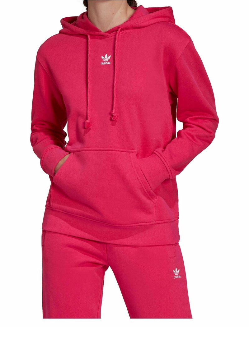 Personne portant un sweat à capuche Adidas rose vif et un pantalon assorti avec les logos trefoil blancs d'Adidas et les mains dans la poche du sweat.
