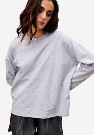 Femme avec de longs cheveux bruns, portant une chemise à manches longues gris clair ample et un pantalon noir, regardant vers le bas avec les mains derrière le dos.