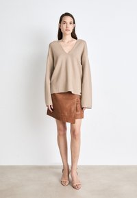 Beige, gestrickter Pullover mit V-Ausschnitt und übergroßen Ärmeln, kombiniert mit einem braunen, ledernen Wickelrock mit einem angenähten Reißverschluss an der Seite.
