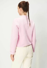 Rosa, texturerad sweatshirt med långa ärmar, kort design och rullade ärmslut, parat med krämfärgade byxor. Bakvy visas.