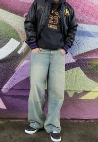 Chaqueta bomber de satén negra con acentos morados, sudadera gris, jeans de mezclilla azul claro holgados y zapatillas negras. Frente a un colorido fondo de graffiti.