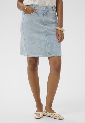 Femme portant une jupe en denim bleu clair à motifs subtils, un haut sans manches crème, des ballerines tressées crème, et des bracelets dorés et beige.