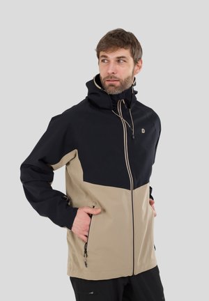Mann trägt eine schwarz-beige Kapuzenjacke mit den Händen in den Taschen und blickt seitlich vor einem einfarbigen grauen Hintergrund.