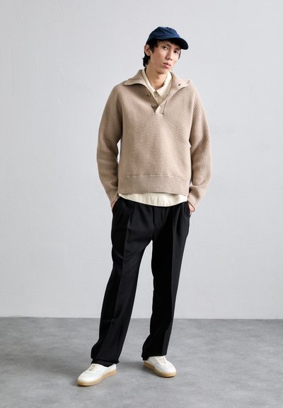 Beige knitted sweater with a collar, worn over a light-colored shirt, paired with black trousers and white sneakers. Navy cap completes the look.