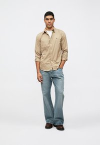 Camicia beige a maniche lunghe con bottoni, jeans blu chiaro a gamba larga, scarpe marroni. Design semplice e casual con una vestibilità comoda.