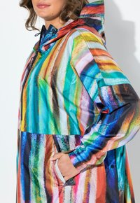 Femme portant un imperméable à rayures multicolores avec fermeture éclair et capuche, main dans la poche, sur fond clair.
