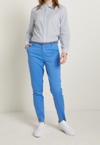 Pantalons ajustés bleus vifs avec une texture lisse, associés à une chemise à rayures légères à manches longues et des baskets blanches, se tenant sur un fond uni.
