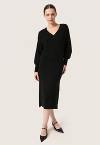 Rochie neagră tricotată cu decolteu în V, model cu torsadă, mâneci lungi și fante laterale. Modelul a asortat-o cu pantofi plata negri cu vârf ascuțit.