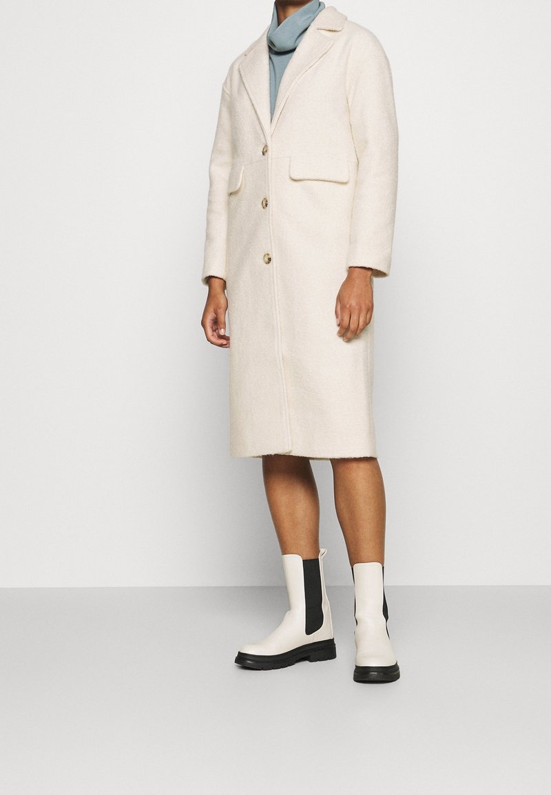 Abrigo de lana beige con cuello, dos bolsillos frontales y tres botones. Combina con botines crema con paneles laterales negros.