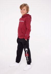 Felpa rossa con cappuccio e tasca frontale, decorata con un logo bianco, abbinata a pantaloni della tuta neri con dettaglio del logo, indossati con sneakers bianche.