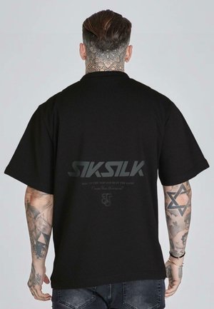 Uomo con tatuaggi sul collo e sul braccio che indossa una maglietta nera oversize SikSilk con logo e scritta sul retro, e jeans scuri.