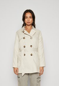 Trench coat bej deschis cu nasturi dubli, talie cu curea și guler. Textură netedă cu două buzunare laterale.