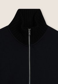 Chaqueta negra con cremallera, que presenta un alto cuello acanalado, textura de tela suave y cierre de cremallera plateada.