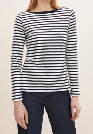 Femme portant un haut blanc à manches longues avec des rayures horizontales noires et un jean bleu foncé, debout devant un fond uni clair.