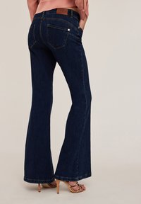 Vaqueros de tiro medio en denim azul oscuro, con campana, dos bolsillos traseros y un parche de cuero marrón en la cinturilla. Textura de denim suave.