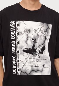 Tricou din bumbac negru, cu un grafic mare monochrom cu un design de vultur, "Versace Jeans Couture" și un motiv de lanț pe latură.