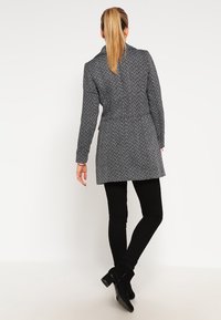 Mujer con coleta rubia que lleva un abrigo gris de espiga, pantalones negros y botines negros, de pie contra un fondo blanco.