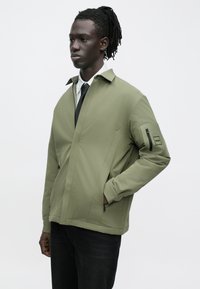 Olive green jacket with a structured design, featuring a stand-up collar, front zip pockets, and a zippered sleeve pocket.
