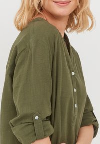 Haut en vert olive texturé avec un col en V, des boutons décoratifs et des manches retroussées. Tissu doux avec une coupe décontractée, idéal pour un look décontracté.