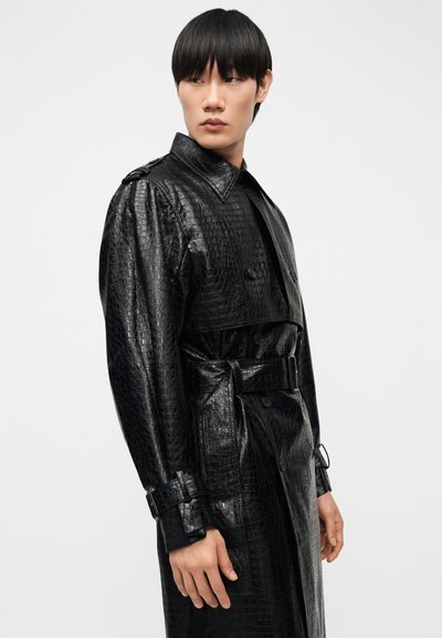 Άτομο που φοράει μαύρο ανάγλυφο δερμάτινο παλτό τύπου τρεντσκότ με ζώνη, κουμπιά και επωμίδες, κοιτάζοντας προς τα πλάγια σε απλό φόντο.