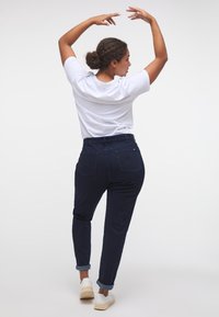 Tumši džinsi ar salocītām piedurknēm, kas izceļ saspringto jostasvietu un divas aizmugurējās kabatas. Kombinēti ar baltu t-kreklu neformālā pozā.