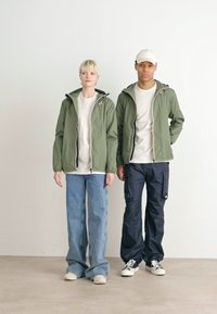 Giubbotti verdi con zip e cappuccio, magliette grigio chiaro, jeans blu larghi e pantaloni cargo neri. Sneakers bianche e un cappello completano gli outfit.