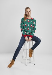 Zöld pulóver Mikulás és hópehely mintákkal, piros díszítéssel, kék farmernadrággal és barna fűzős csizmával párosítva, fehér széken ülve.