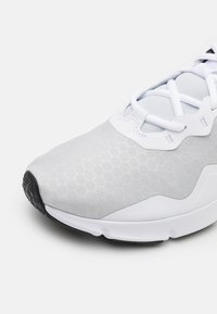 Jasnoszary but sportowy z teksturowanym wzorem heksagonalnym, białymi akcentami, sznurowanym zapięciem oraz czarną gumową podeszwą.